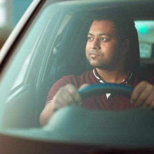 Portrait of an Asian/ Indian man driving a car, holding string and looking in front of camera in city street on a dusky evening at sunset time.