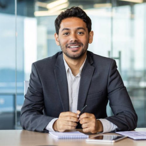 Portrait of young hispanic businessman inside office, boss in business suit smiling and looking at camera, experienced satisfied man at workplace at desk.